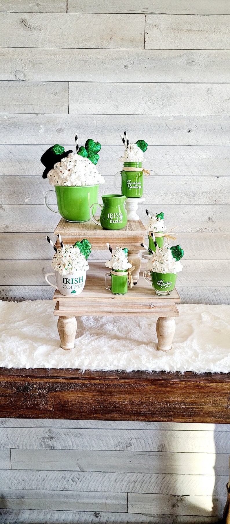 May include: A tiered wooden stand with four green and white mugs with shamrock designs and the text "Irish Brew", "Irish Coffee", and "Lucky". The mugs are topped with whipped cream and green glitter.