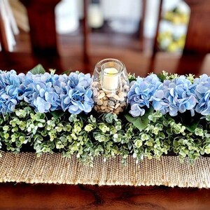 May include: A blue and green floral centerpiece with a glass candle holder filled with white stones. The centerpiece is on a brown table with a burlap runner.
