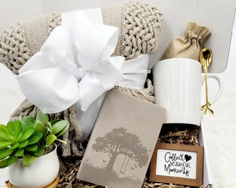 May include: A gift box filled with items for self-care and relaxation. The box includes a white mug, a small bag of tea, a journal with a tree and swing design, a small potted succulent plant, a throw blanket, and a sign that reads "Collect beautiful moments".