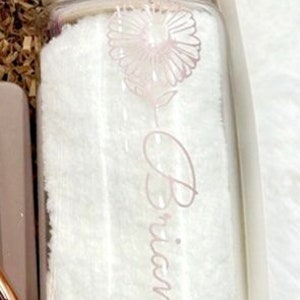 May include: A clear glass jar with a wooden lid and a straw. The jar is filled with a white fluffy material and has a pink flower design and the name "Brianna" written on it.