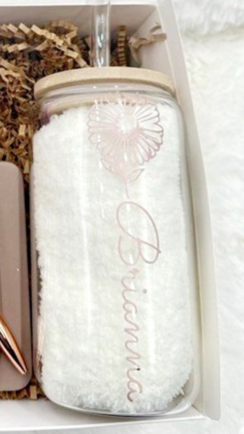 May include: A clear glass jar with a wooden lid and a straw. The jar is filled with a white fluffy material and has a pink flower design and the name "Brianna" written on it.