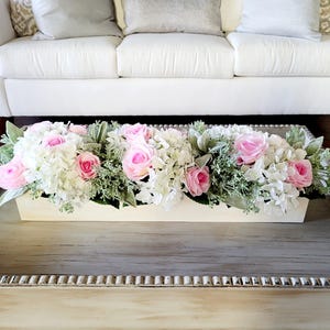 May include: A white wooden planter box filled with a floral arrangement of pink roses and white hydrangeas. The planter is sitting on a gray wooden coffee table.