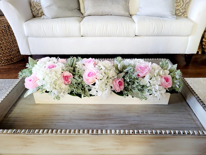 May include: A white wooden planter box filled with a floral arrangement of pink roses and white hydrangeas. The planter is sitting on a gray wooden coffee table.