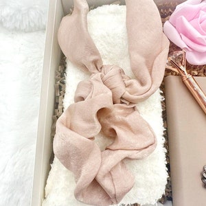 May include: A gift box filled with items for self-care. The box contains a white fuzzy blanket, a pink satin hair scrunchie, a pink rose, and a gold pen with a crystal top.