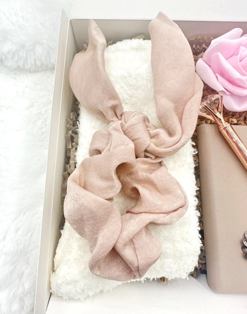 May include: A gift box filled with items for self-care. The box contains a white fuzzy blanket, a pink satin hair scrunchie, a pink rose, and a gold pen with a crystal top.