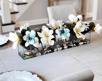 May include: A rectangular glass vase filled with artificial magnolia flowers featuring white and light blue petals, dark green leaves, and small pieces of driftwood arranged along the bottom of the clear container to create a floral centerpiece.