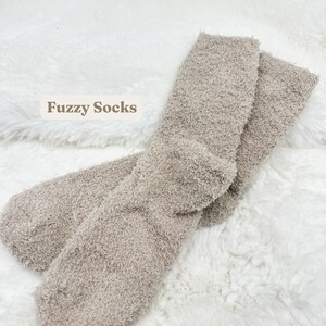May include: A pair of soft, fuzzy socks in a neutral taupe color. The socks are displayed on a white, fluffy surface. The text "Fuzzy Socks" is visible in the image.