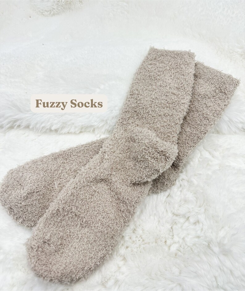 May include: A pair of soft, fuzzy socks in a neutral taupe color. The socks are displayed on a white, fluffy surface. The text "Fuzzy Socks" is visible in the image.