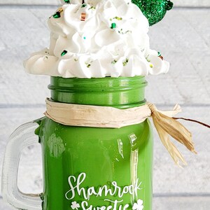 May include: Green glass mason jar with a white handle and a white shamrock design that says "Shamrock Sweetie". The jar is filled with white whipped cream and green glitter. There is a green glitter shamrock on top of the whipped cream. The jar is sitting on a white pedestal.