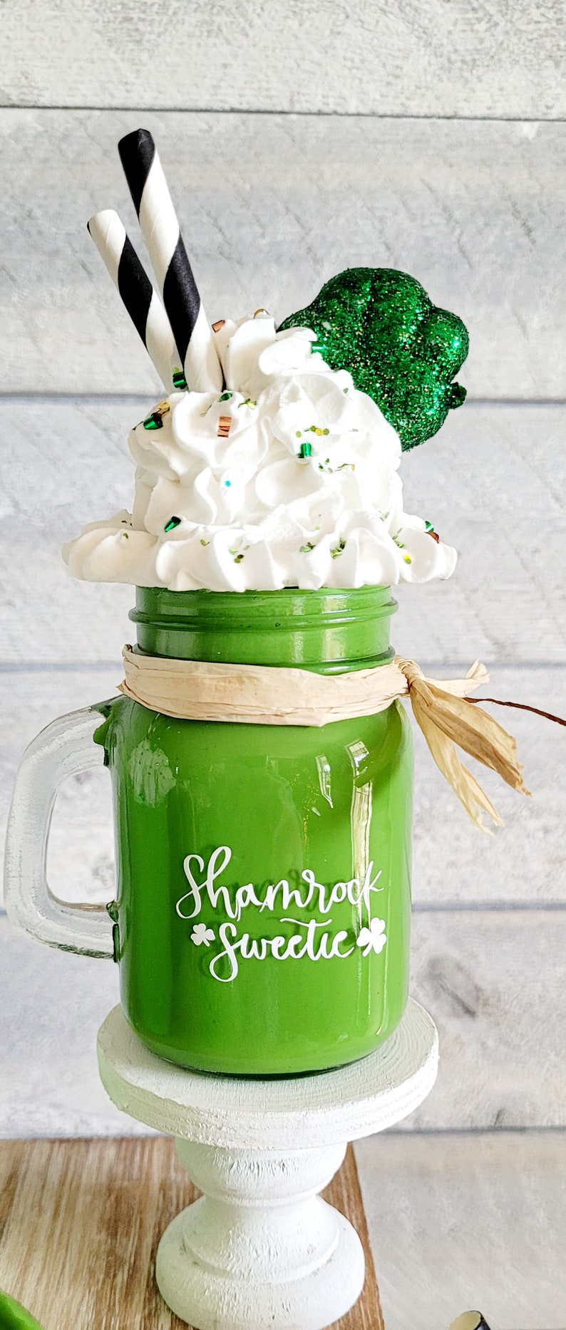 May include: Green glass mason jar with a white handle and a white shamrock design that says "Shamrock Sweetie". The jar is filled with white whipped cream and green glitter. There is a green glitter shamrock on top of the whipped cream. The jar is sitting on a white pedestal.