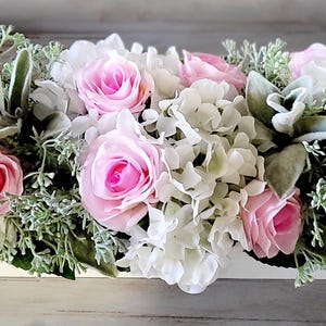 May include: A white wooden planter filled with a floral arrangement of pink roses and white hydrangeas. The arrangement is accented with green foliage.