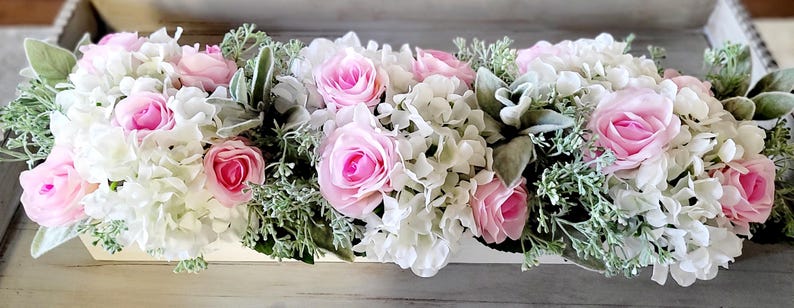 May include: A white wooden planter filled with a floral arrangement of pink roses and white hydrangeas. The arrangement is accented with green foliage.