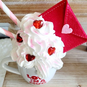 May include: A white mug with the words "Love in a Mug" printed on it. The mug is filled with whipped cream and topped with red heart-shaped sprinkles. A red felt envelope with a pink heart is on top of the whipped cream.