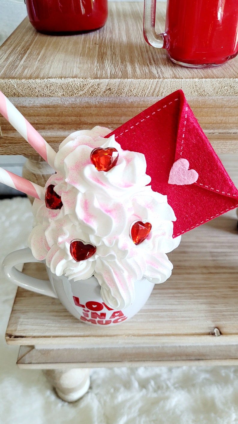 May include: A white mug with the words "Love in a Mug" printed on it. The mug is filled with whipped cream and topped with red heart-shaped sprinkles. A red felt envelope with a pink heart is on top of the whipped cream.