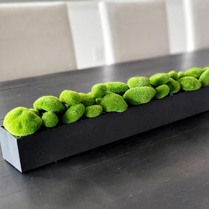 Może przedstawiać: Długi, czarny pojemnik na rośliny wypełniony zielonymi kamieniami mchu. Pojemnik stoi na ciemnym drewnianym stole.