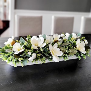 May include: A rectangular white floral arrangement with large white magnolia flowers, small white blooms, and green leaves. The display sits on a dark wooden table, providing a striking contrast.