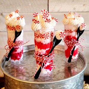 May include: Three red and white peppermint-themed desserts in clear glass cups with red and white string and a black spoon. The desserts are on a silver metal surface.