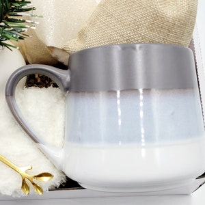 May include: A gray, white, and blue ceramic mug with a rounded shape and a handle. The mug is sitting on a white surface.
