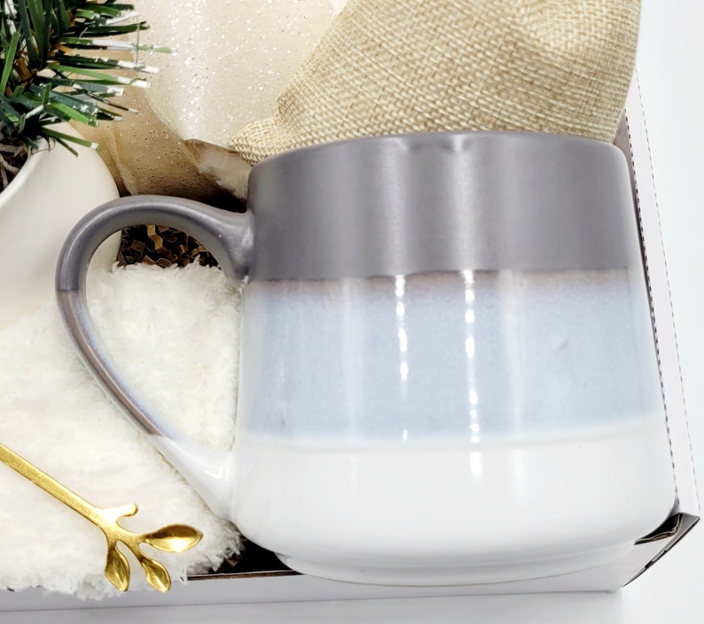 May include: A gray, white, and blue ceramic mug with a rounded shape and a handle. The mug is sitting on a white surface.