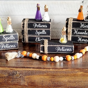 May include: Four black wooden boxes with white text, stacked on top of each other, with small glass bottles filled with colorful liquids on top. The boxes are labeled "Potions Spells Elixirs", "Spells Potions", "Hicks Haunted House", and "Potions". A black and orange beaded garland with a brown tassel is in the foreground.