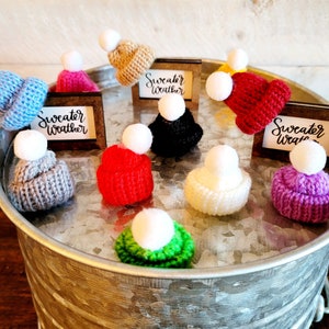 Puede incluir: Surtido de diez gorros de punto con pompones en varios colores, incluyendo rojo, azul, verde, morado, blanco, gris y marr&oacute;n. Los gorros se muestran en una bandeja de metal plateado con el texto "Sweater Weather" en tres peque&ntilde;as placas de madera.