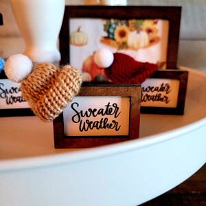 Puede incluir: Una bandeja de tres niveles con tres peque&ntilde;as se&ntilde;ales enmarcadas. Las se&ntilde;ales tienen el texto "Sweater Weather". La bandeja est&aacute; decorada con un peque&ntilde;o gorro de punto con un pomp&oacute;n blanco y un peque&ntilde;o gorro rojo de punto con un pomp&oacute;n blanco.