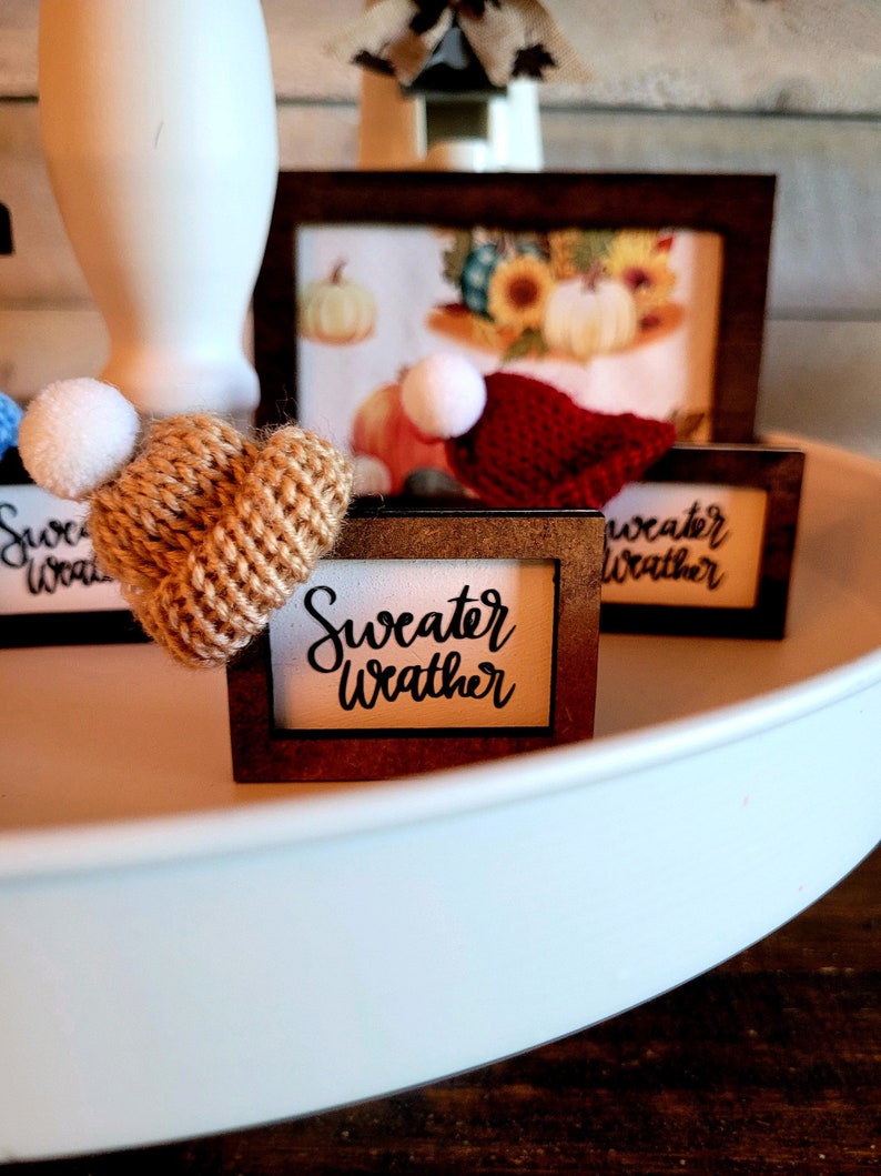 Puede incluir: Una bandeja de tres niveles con tres peque&ntilde;as se&ntilde;ales enmarcadas. Las se&ntilde;ales tienen el texto "Sweater Weather". La bandeja est&aacute; decorada con un peque&ntilde;o gorro de punto con un pomp&oacute;n blanco y un peque&ntilde;o gorro rojo de punto con un pomp&oacute;n blanco.