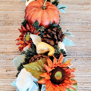 May include: A fall-themed centerpiece with a small orange pumpkin, white gourds, pine cones, sunflowers, and white flowers on a brown woven placemat.