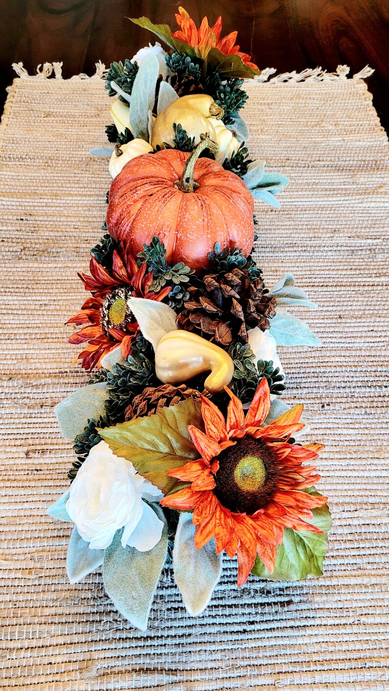 May include: A fall-themed centerpiece with a small orange pumpkin, white gourds, pine cones, sunflowers, and white flowers on a brown woven placemat.