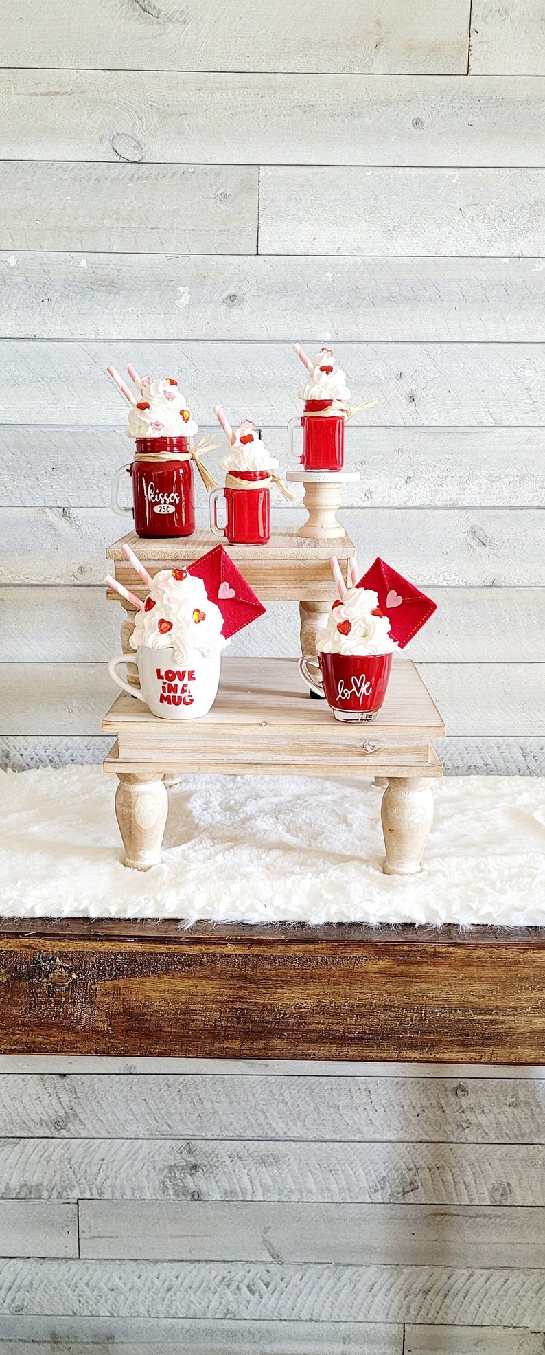 May include: Four mugs with whipped cream and red hearts on top, sitting on two wooden tiered stands. The mugs are decorated with the words "Love in a Mug" and "Kisses 25c".