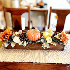 May include: A wooden box filled with fall-themed decor, including a small orange pumpkin, yellow gourds, pine cones, and sunflowers. The box is sitting on a woven rug.