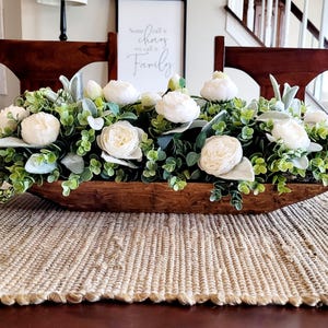 May include: A wooden dough bowl filled with white flowers and greenery. The bowl is sitting on a woven placemat on a wooden table. The text "All is changing, we call it a family" is visible on a framed print in the background.