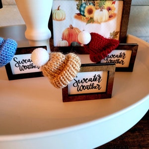 Puede incluir: Tres peque&ntilde;as se&ntilde;ales de madera con el texto "sweater weather" escrito en cursiva negra. Cada se&ntilde;al tiene un peque&ntilde;o gorro de punto en la parte superior, uno azul, uno marr&oacute;n y uno rojo.