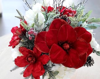 May include: A floral arrangement featuring large red velvet magnolia blossoms with glittered edges, white flowers, frosted pine branches, small red berries, and dark pinecones. The composition includes thin, bare, dark twigs extending upwards.