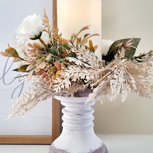 Op de afbeelding: Een witte pilaar-kaarshouder met een bruine houten basis. De kaarshouder is versierd met een krans van gedroogde bloemen en groen, waaronder witte rozen, beige pampasgras en bruine takjes.