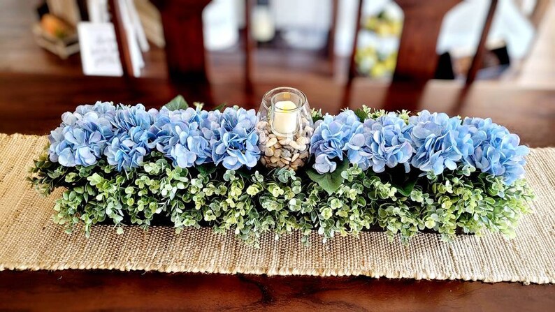 May include: A blue and green floral centerpiece with a glass candle holder filled with white stones. The centerpiece is on a brown table with a burlap runner.