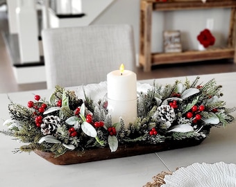 May include: A rustic wooden dough bowl centerpiece filled with artificial evergreen branches, frosted pinecones, and bright red berries. A single white pillar candle sits in a clear glass holder at the center of the arrangement, creating a festive display.