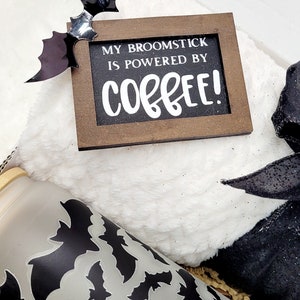 Puede incluir: Una caja de regalo con tem&aacute;tica de Halloween con una manta blanca peluda, una cinta negra y blanca, un vaso transparente con un patr&oacute;n de murci&eacute;lago negro y un letrero de madera que dice "My broomstick is powered by coffee!"