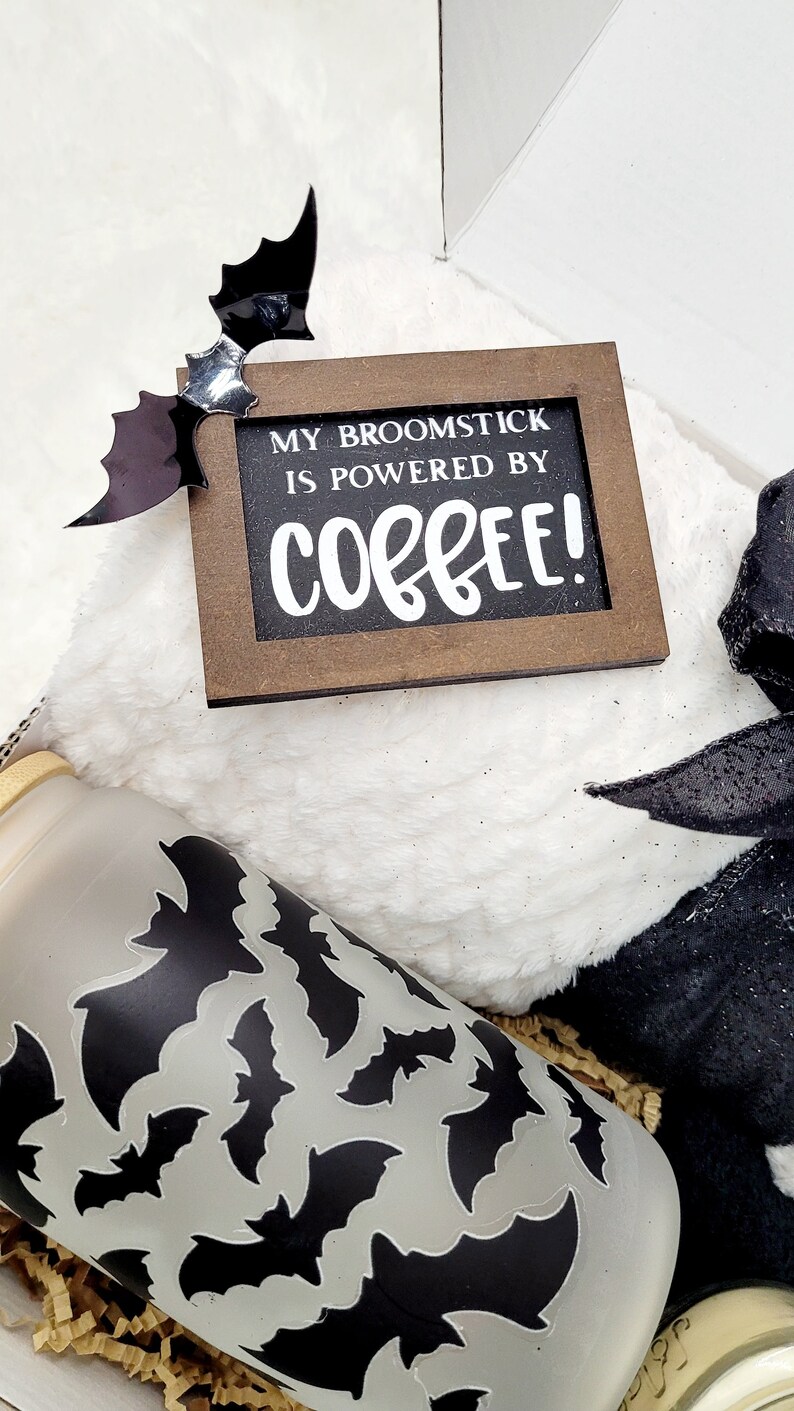 Puede incluir: Una caja de regalo con tem&aacute;tica de Halloween con una manta blanca peluda, una cinta negra y blanca, un vaso transparente con un patr&oacute;n de murci&eacute;lago negro y un letrero de madera que dice "My broomstick is powered by coffee!"