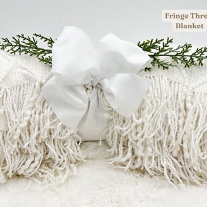 May include: A folded white fringe throw blanket with a large white bow and sprigs of greenery. The blanket has a textured weave and long fringe along the edges. Text on the image reads "Fringe Throw Blanket."