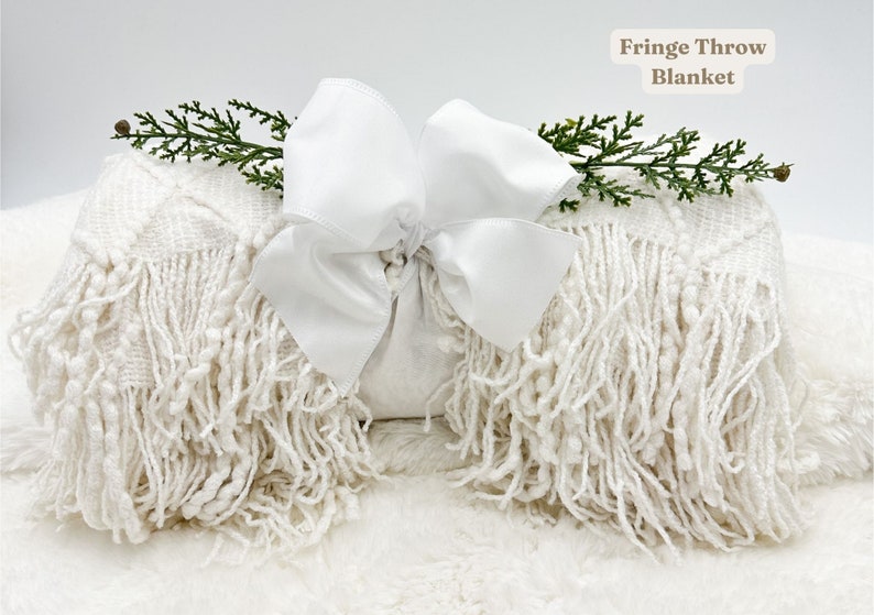 May include: A folded white fringe throw blanket with a large white bow and sprigs of greenery. The blanket has a textured weave and long fringe along the edges. Text on the image reads "Fringe Throw Blanket."