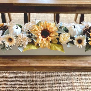 May include: A rectangular white floral arrangement featuring sunflowers, white pumpkins, and various fall-coloured flowers and greenery. The arrangement is placed on a wooden table, creating a seasonal centrepiece.
