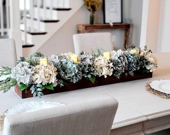 May include: A long, rectangular dark brown wooden box centerpiece filled with artificial cream and blue hydrangea flowers, green foliage, and three clear glass candle holders containing glowing yellow LED pillar candles.