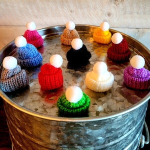 Puede incluir: Una colecci&oacute;n de 11 gorros de punto de colores con pompones blancos. Los gorros est&aacute;n dispuestos en c&iacute;rculo sobre una bandeja de metal plateado.