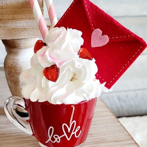 May include: A red ceramic mug with a white "love" design, filled with whipped cream and red heart sprinkles. A red felt envelope with a pink heart is on top of the whipped cream. Two pink and white striped straws are sticking out of the whipped cream.