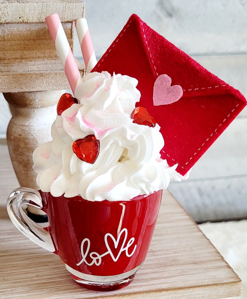 May include: A red ceramic mug with a white "love" design, filled with whipped cream and red heart sprinkles. A red felt envelope with a pink heart is on top of the whipped cream. Two pink and white striped straws are sticking out of the whipped cream.