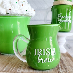 May include: A green glass creamer with a clear handle. The creamer has the words "Irish Brew" written on the front in white.
