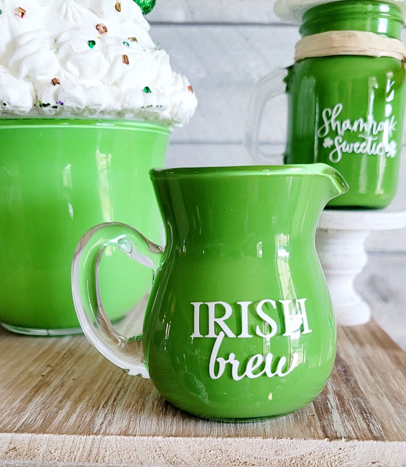 May include: A green glass creamer with a clear handle. The creamer has the words "Irish Brew" written on the front in white.