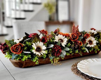 May include: A long, rectangular, dark brown wooden dough bowl filled with an arrangement of artificial flowers and greenery, including cream-colored sunflowers, deep orange roses, dark red blossoms, and small green leaves.