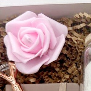May include: A pink rose, a gold-colored pen with a crystal-like top, and a glass jar filled with bath salts. The items are arranged in a white gift box with brown shredded paper.